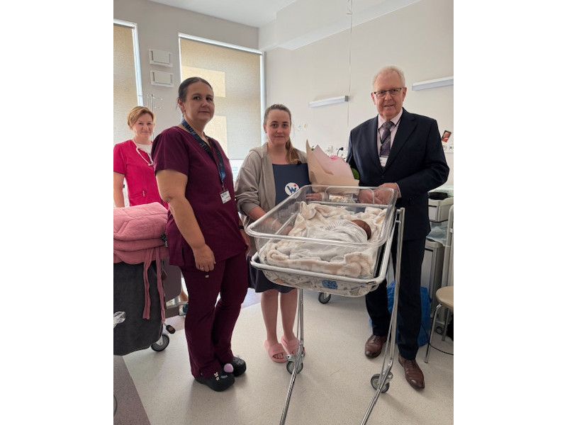 Sali poporodow Wojewódzkiego Szpitala Specjalistycznego im. Stefana Kardynała Wyszyńskiego w Lublinie stoją razem cztery osoby: od prawej strony dyrektor szpitala Piotr Matej, po środku matka dziecka stojąca za przezroczystym łóżeczkiem z przykrytym 1000 noworodkiem, po lewej 2 pielęgniarki w kolorowych uniformach.