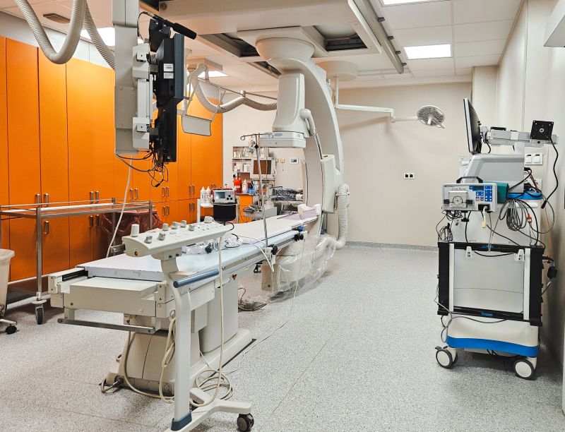Nowocześnie wyposażona sala zabiegowa kardiologii inwazyjnej z długim stołem zabiegowym pośrodku, nad którym znajduje się łuk aparatu angiograficznego z monitorami i przewodami; po lewej stronie widać rząd pomarańczowych szaf zabudowanych, a po prawej mobilny wózek z aparaturą medyczną i ekranem, całość utrzymana jest w jasnych, sterylnych barwach.