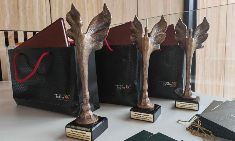 Na jasnym stole ustawione są trzy metalowe statuetki z figurą stylizowanego anioła o uniesionych skrzydłach, umieszczone na czarnych postumentach z napisem „Pracodawca Roku 2024”, a za nimi stoją czarne torby prezentowe z czerwonymi uchwytami i logo „Lublin – Miasto Inspiracji”; obok widoczne są eleganckie teczki i dyplomy przygotowane na uroczystą galę wręczenia nagród.