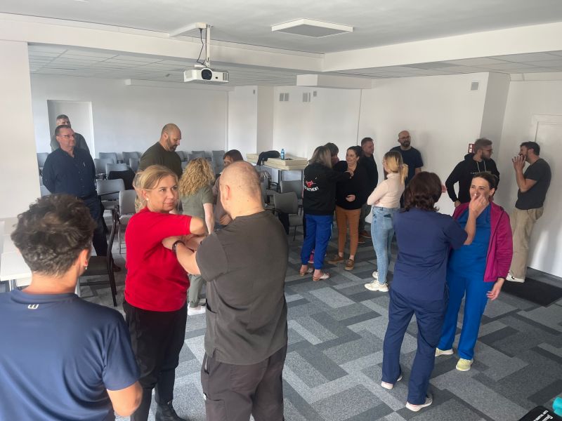 grupa pracowników szpitala uczestniczy w praktycznych ćwiczeniach samoobrony i deeskalacji agresji. Uczestnicy, ustawieni w parach lub małych grupach, ćwiczą techniki pod okiem instruktora, w jasno oświetlonym pomieszczeniu z rzędami krzeseł w tle.