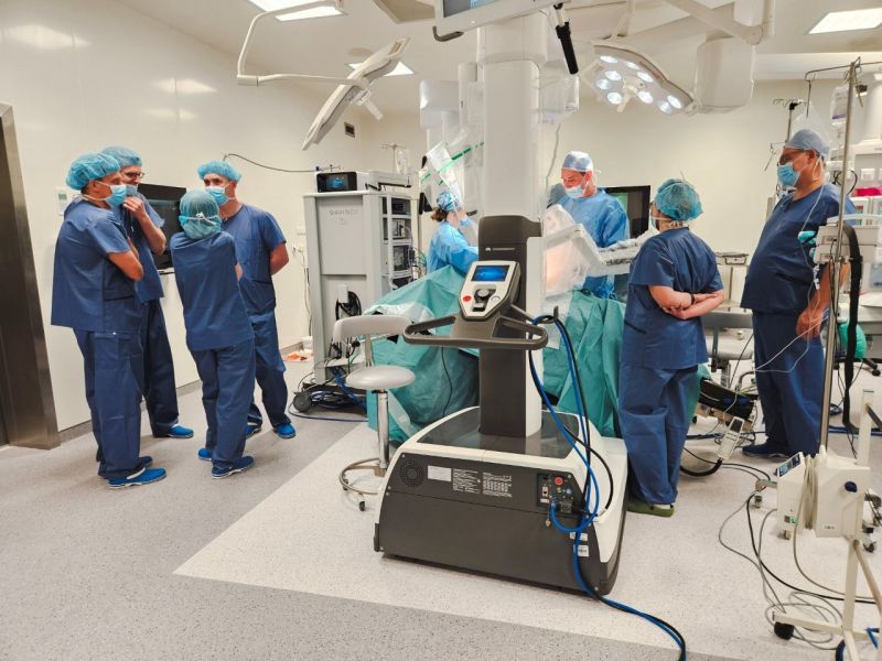 Na sali operacyjnej przy robocie chirurgicznym da Vinci pracuje zespół ubranych na niebiesko chirurgów, podczas gdy inni uczestnicy warsztatów stoją obok w grupach i omawiają przebieg zabiegu; wokół stołu operacyjnego widoczna jest nowoczesna aparatura medyczna, monitory oraz osłonięte zielonymi serwetami pole operacyjne.