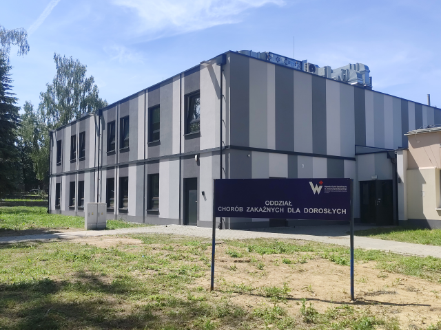 Zdjęcie przedstawia nowoczesny, dwukondygnacyjny budynek Oddziału Chorób Zakaźnych dla Dorosłych, o elewacji w pionowe pasy w odcieniach szarości. W pierwszym planie znajduje się duża, ciemnoniebieska tablica informacyjna z wyraźnym, białym napisem: "ODDZIAŁ CHORÓB ZAKAŹNYCH DLA DOROSŁYCH" oraz logo szpitala. Budynek otoczony jest trawnikiem i wysokimi drzewami pod błękitnym niebem.