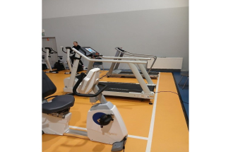 Sala ćwiczeń, na pierwszym planie znajduje się rowerek stacionarny i dwie bierznie, wszystko stoi na pomarańczowej podłodze sportowej. W tle widać ćwiczącą osobę oraz kilka innych sprzętów fitness przy szarej ścianie.