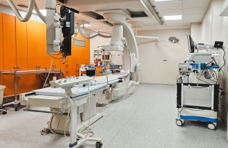 Nowocześnie wyposażona sala zabiegowa kardiologii inwazyjnej z długim stołem zabiegowym pośrodku, nad którym znajduje się łuk aparatu angiograficznego z monitorami i przewodami; po lewej stronie widać rząd pomarańczowych szaf zabudowanych, a po prawej mobilny wózek z aparaturą medyczną i ekranem, całość utrzymana jest w jasnych, sterylnych barwach.