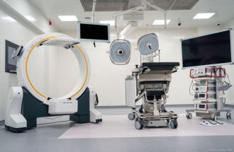 Nowocześnie wyposażona sala operacyjna z dużym, okrągłym ramieniem aparatu obrazującego po lewej stronie, pośrodku z mobilnym stołem operacyjnym i dwiema lampami operacyjnymi podwieszonymi do sufitu oraz po prawej z wysokim monitorem i wózkiem z aparaturą medyczną, w jasnym, sterylnym wnętrzu szpitalnym.