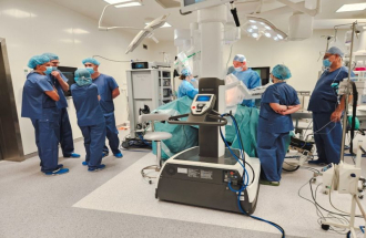 Na sali operacyjnej przy robocie chirurgicznym da Vinci pracuje zespół ubranych na niebiesko chirurgów, podczas gdy inni uczestnicy warsztatów stoją obok w grupach i omawiają przebieg zabiegu; wokół stołu operacyjnego widoczna jest nowoczesna aparatura medyczna, monitory oraz osłonięte zielonymi serwetami pole operacyjne.