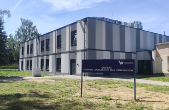 Zdjęcie przedstawia nowoczesny, dwukondygnacyjny budynek Oddziału Chorób Zakaźnych dla Dorosłych, o elewacji w pionowe pasy w odcieniach szarości. W pierwszym planie znajduje się duża, ciemnoniebieska tablica informacyjna z wyraźnym, białym napisem: 