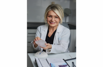 Zdjęcie profesor doktor habilitowanej nauk medycznych Barbary Madej-Czerwonki siedzącej przy biurku w gabinecie lekarskim. Lekarka uśmiecha się do aparatu, ubrana w biały fartuch i okulary, i trzyma w dłoniach silikonowy implant piersi. Zdjęcie wykonane z okazji 15-lecia chirurgii piersi.